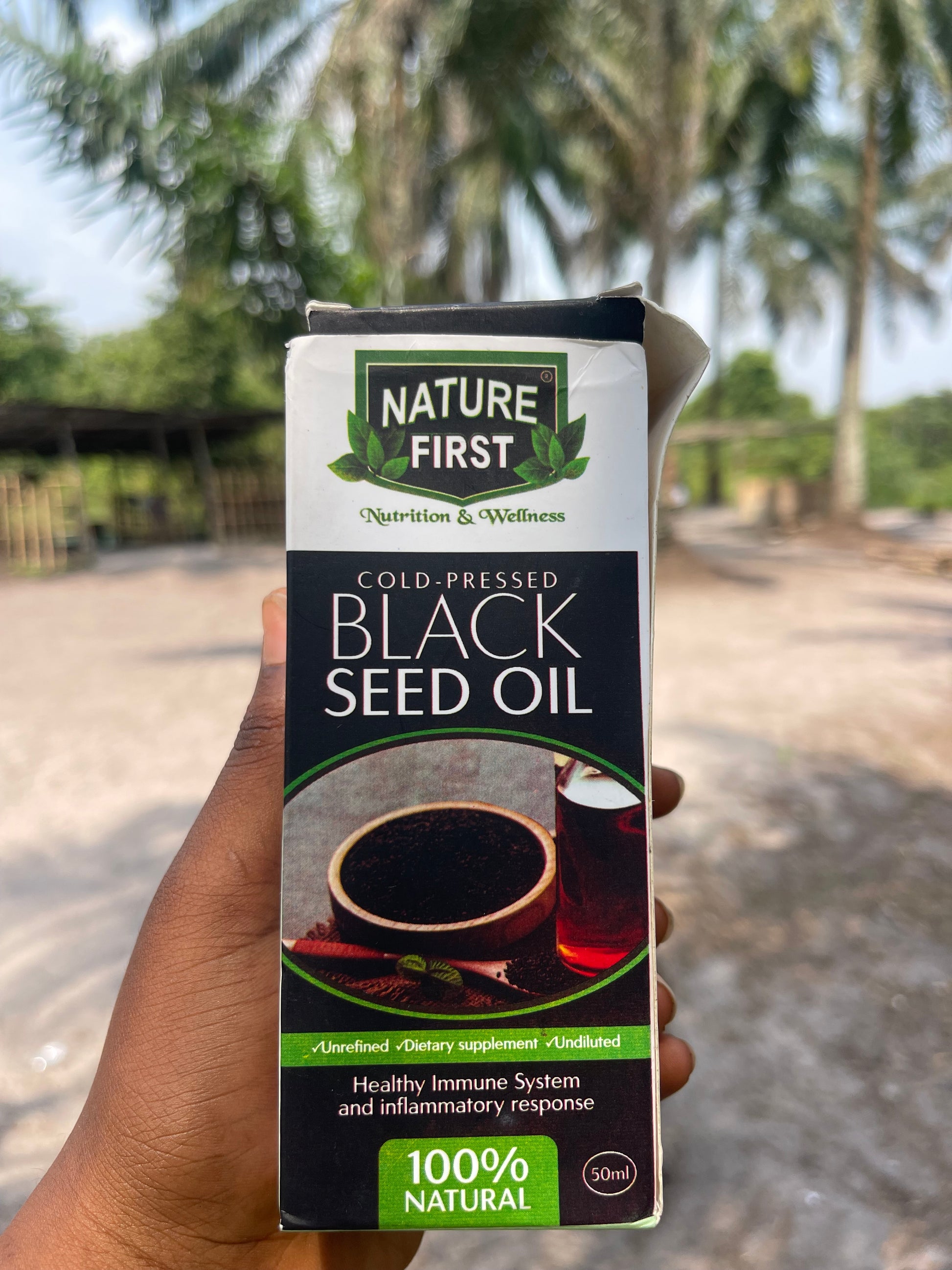 Hand holding a box of Nature First Cold Pressed Black Seed Oil with a blurred natural background.